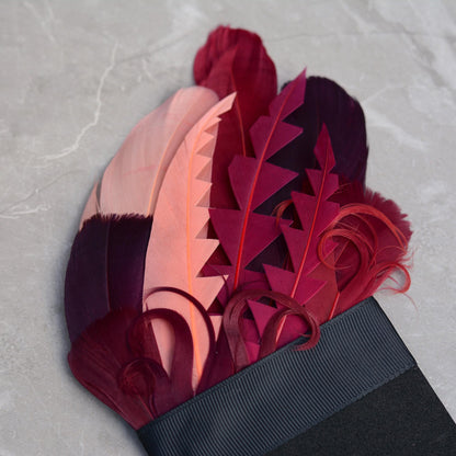 A close-up of a decorative feather suit accessory. A feather pocket square featuring layered goose feathers in deep burgundy, magenta, coral pink, and dark plum tones, cut into graphic shapes and curled for texture and depth.