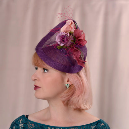 Woman with a bob in rose pink wearing a purple side perching royal ascot  fascinator hat decorated with purple and pink flowers and pink veiling. It is a suitable size to conform with royal ascots royal enclosure rules.