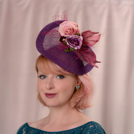 Woman with a bob in rose pink wearing a purple side perching fascinator hat decorated with purple and pink flowers and pink veiling