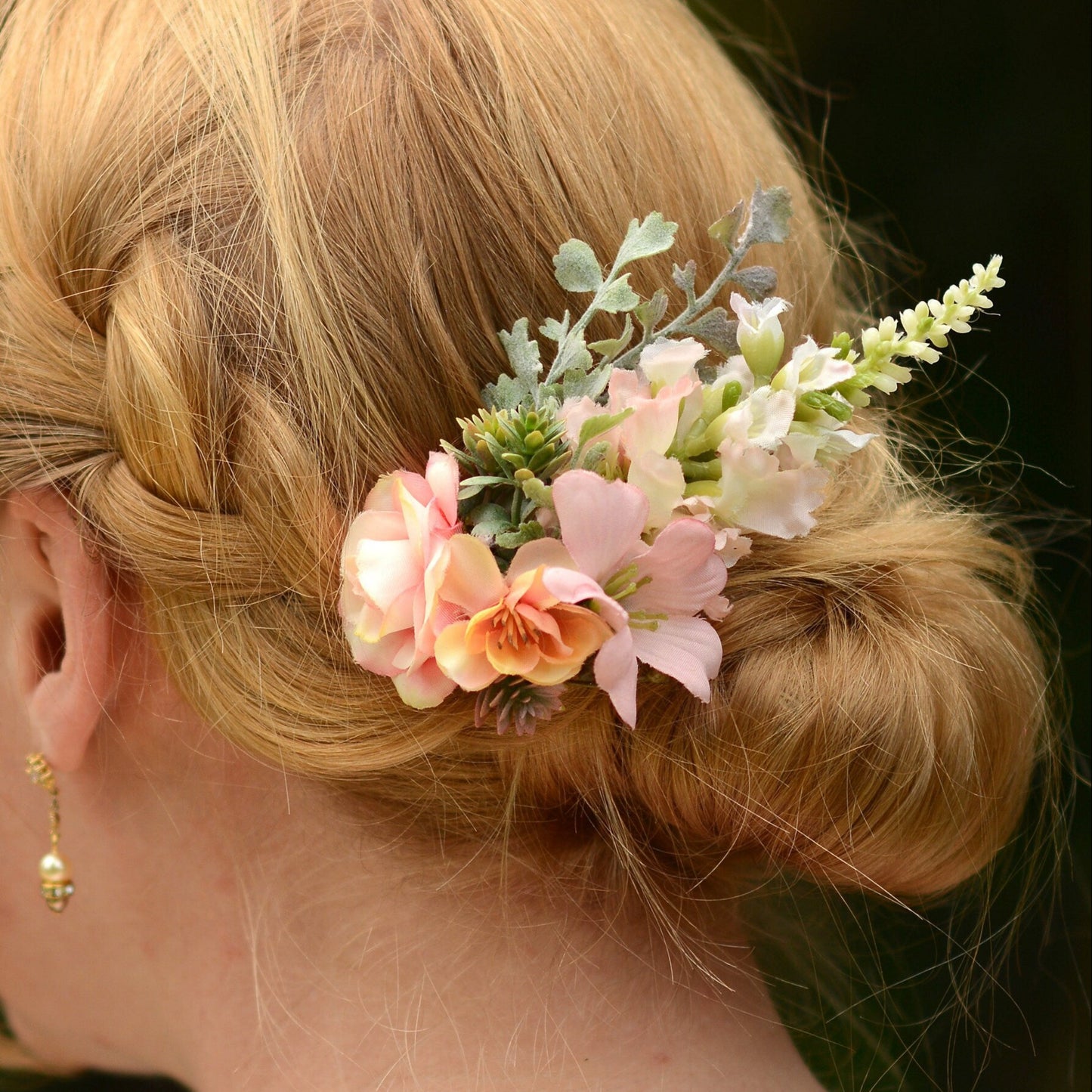 A close-up of a blonde woman's braided bun hairstyle decorated with a delicate floral hair accessory featuring pink and peach flowers, green foliage, and small white blossoms.