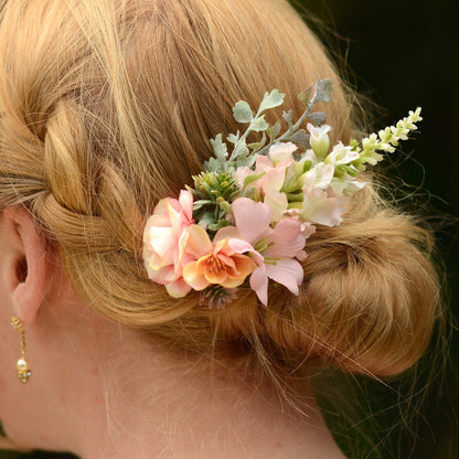 A close-up of a blonde woman's braided bun hairstyle decorated with a delicate floral hair accessory featuring pink and peach flowers, green foliage, and small white blossoms.