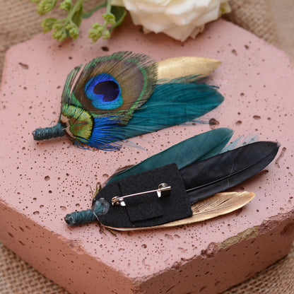 Two decorative feather pin-back brooch accessories displayed on a pink stone slab. One piece features a peacock feather with blue, green, and gold tones, and the other has teal, black, and gold feathers attached to a black fabric backing with a silver safety pin.