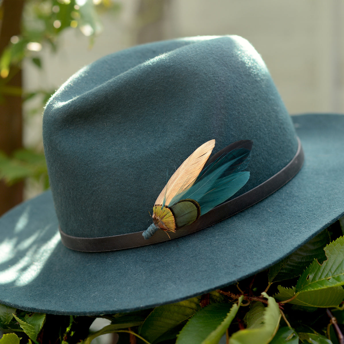 A wide-brimmed felt hat in muted teal-blue with a dark band and a decorative feather hat pin embellishment in teal, green and gold, photographed outdoors among green leaves.