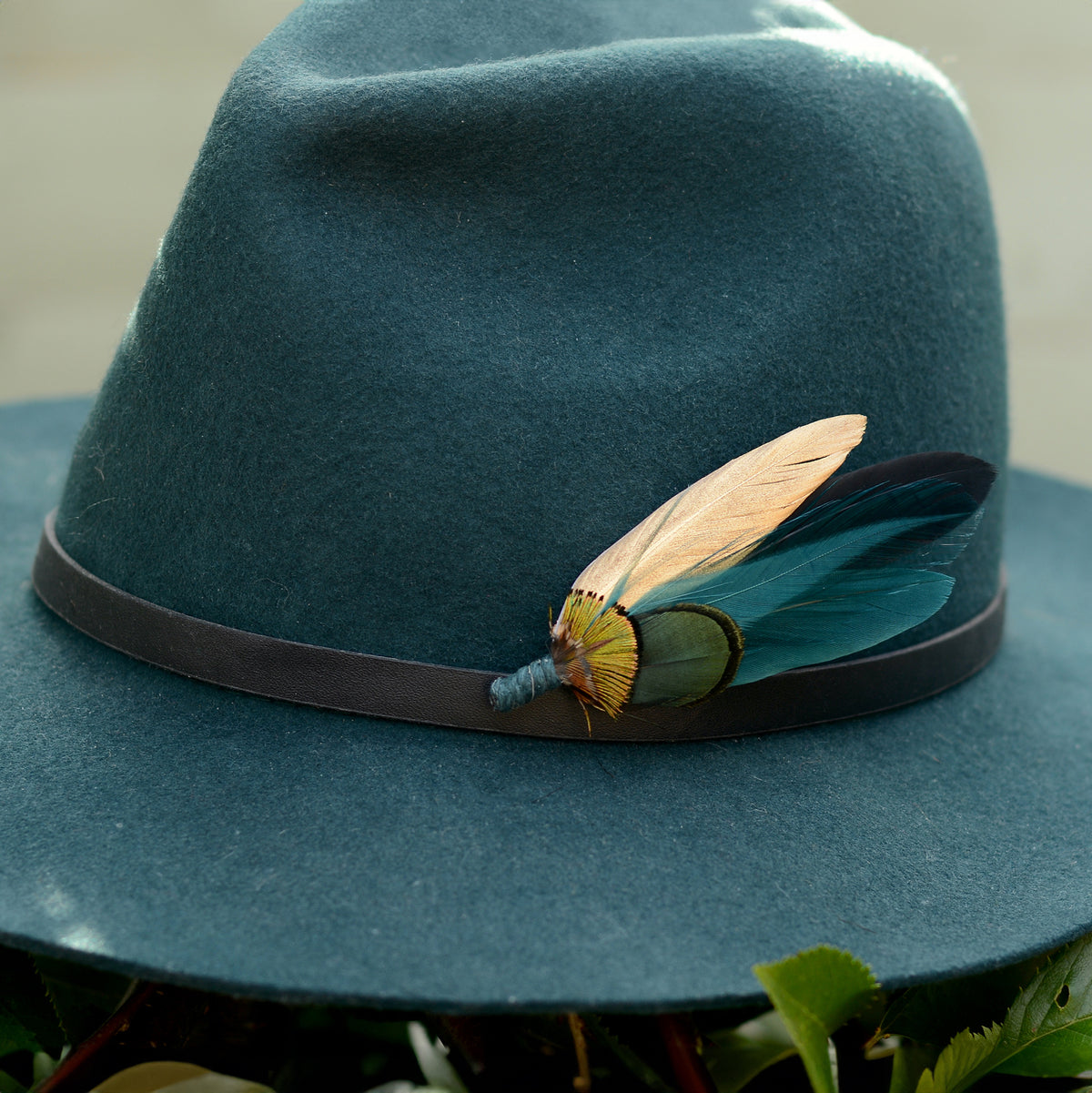 A close up of a wide-brimmed felt hat in muted teal-blue with a dark band and a decorative feather hat pin embellishment in teal, green and gold, photographed outdoors among green leaves.