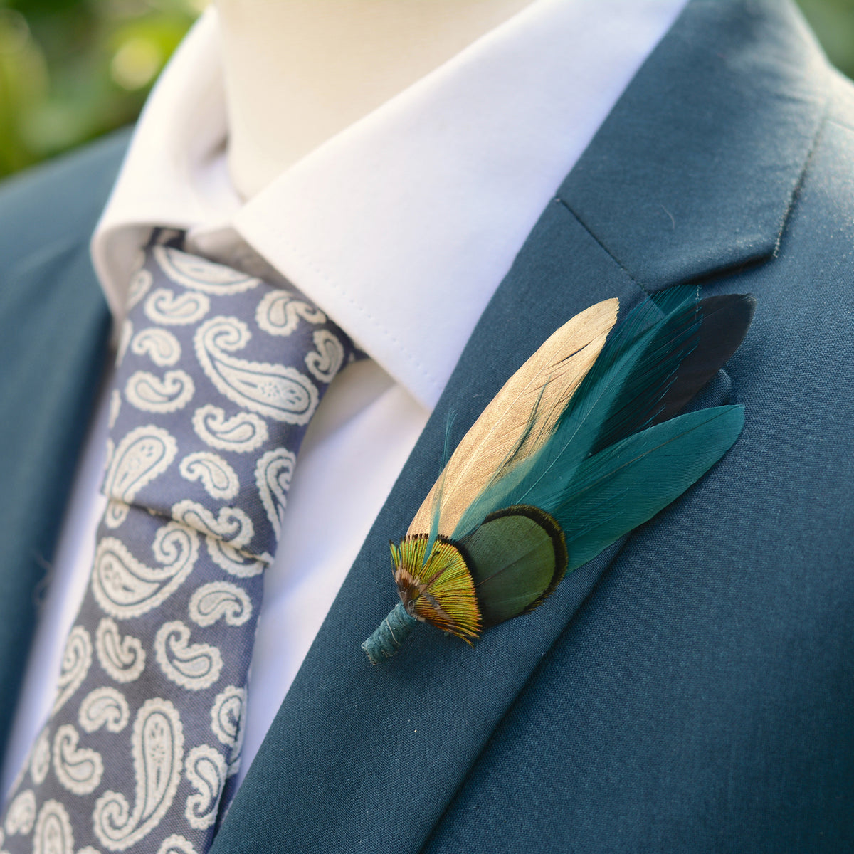 Teal, green and gold feather groomsmen boutonniere on a teal suit jacket surounded by greenery.