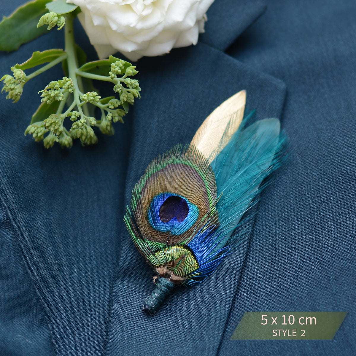 A peacock feather boutonniere pin with vibrant blue, green, and gold eye spots, pinned to a dark navy blue suit jacket lapel next to a white rose stem. A green label in the bottom right corner reads '5 x 10 cm STYLE 2'.