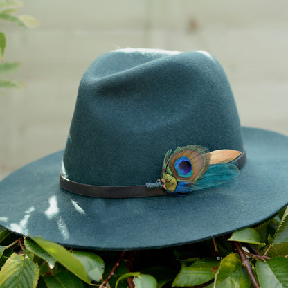 A dark teal felt wide-brim hat resting on green leaves, featuring a black band and a decorative peacock feather hat pin accent.