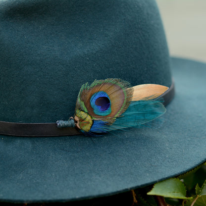 A dark teal felt hat with a black band, decorated with a vibrant peacock feather hat pin showing blue, green, and gold tones.