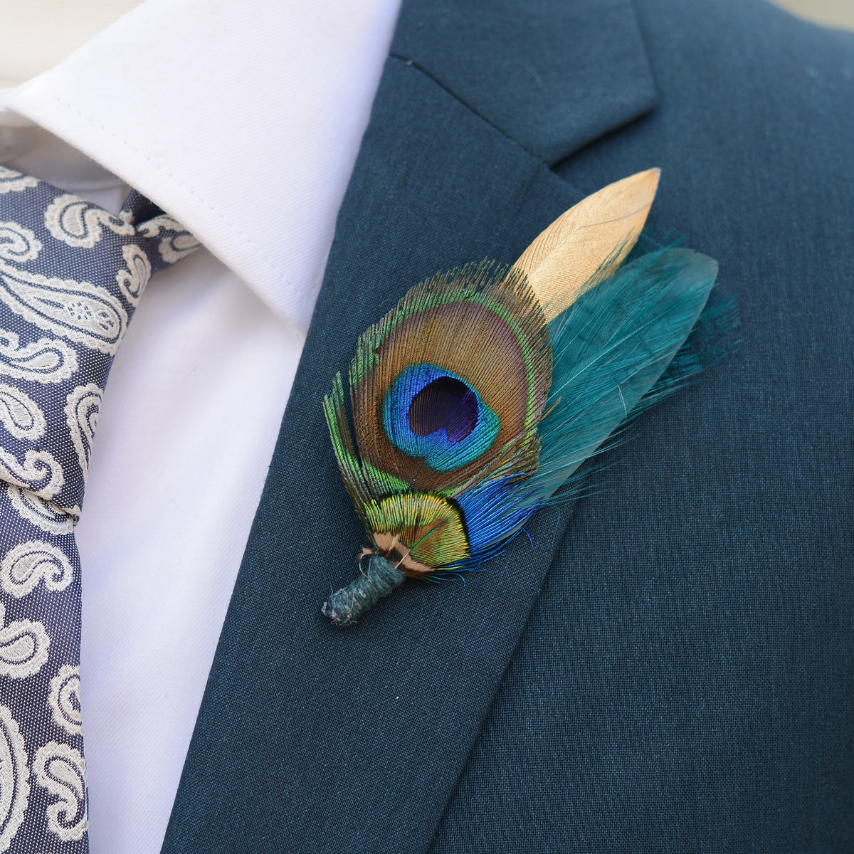 A close-up of a men's formal outfit featuring a dark teal suit jacket, a white dress shirt, a patterned blue and white tie, and a decorative peacock feather boutonniere with teal and gold feathers on the lapel.