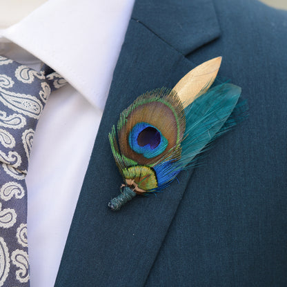A close-up of a men's formal outfit featuring a dark teal suit jacket, a white dress shirt, a patterned blue and white tie, and a decorative peacock feather boutonniere with teal and gold feathers on the lapel.