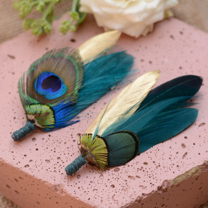 Two decorative peacock feather lapel pins with gold, teal, and blue feathers, displayed on a textured pink block with a white rose in the background.
