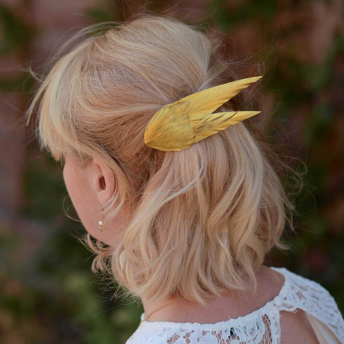 A blonde woman shown from the back wearing a large, textured golden feather bridal hair clip in her hair, with a soft-focus outdoor background.