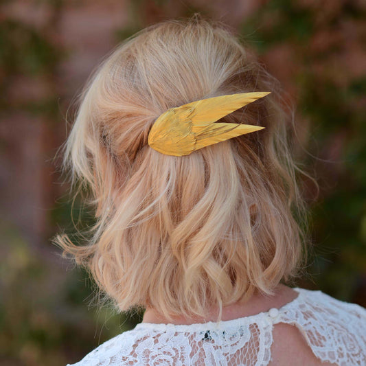 A blonde woman viewed from the back, wearing a metallic gold feather hair clip in her hair, with a white lace top and a blurred outdoor background.