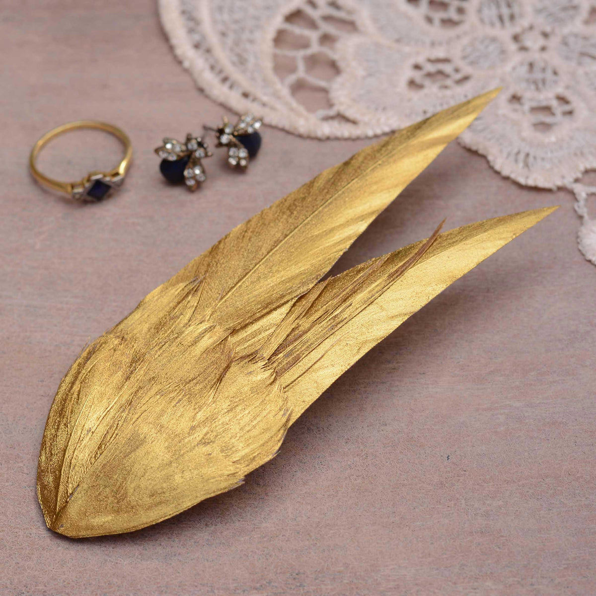 A feather hair fascinator hand painted in warm metallic gold with a elegant v shape. Displayed on a wooden surface with jewellery and lace in the background.