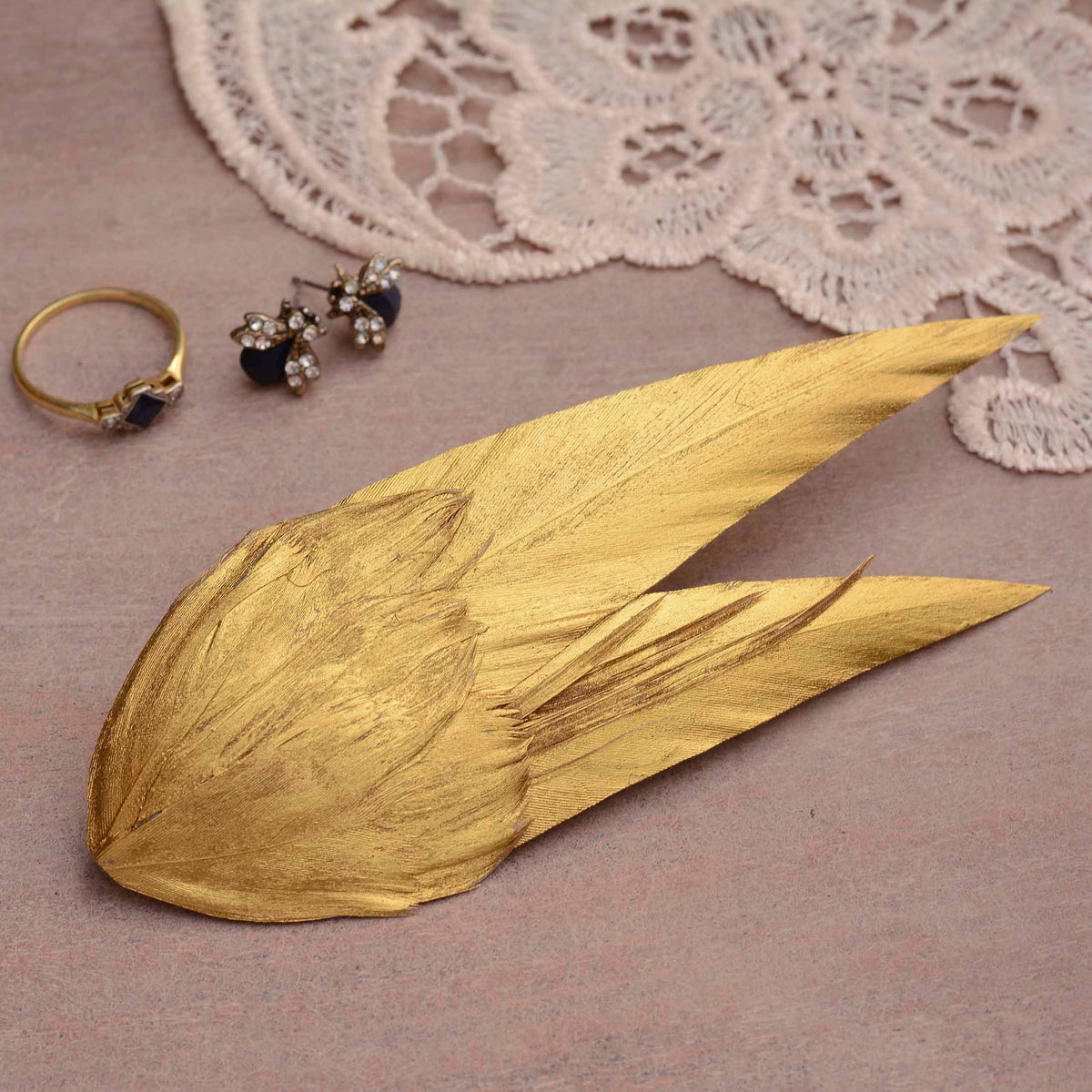 A bridal feather hair accessory hand painted in warm metallic gold with a elegant v shape. Displayed on a wooden surface with jewellery and lace in the background.