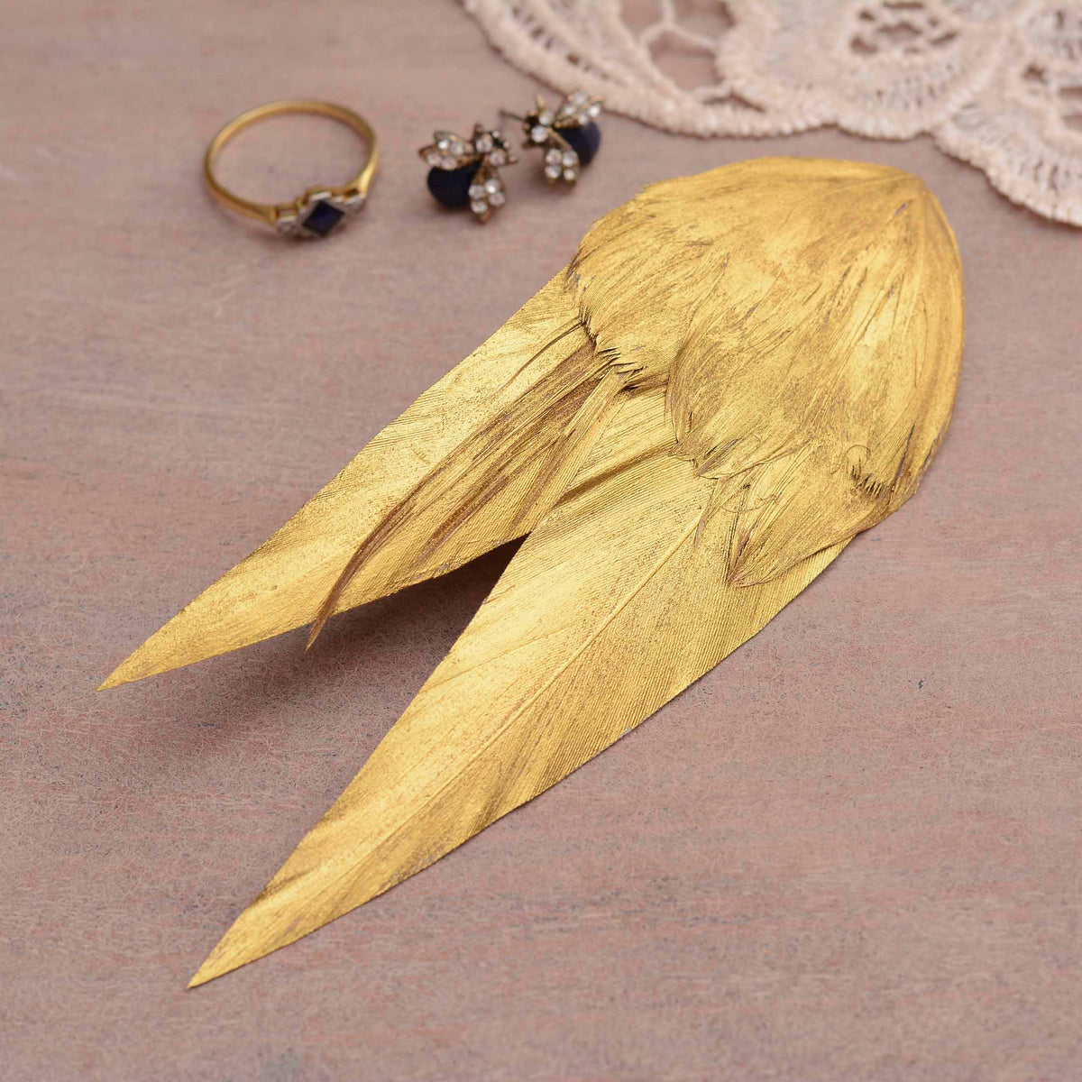 A decorative feather hair accessory hand painted in warm metallic gold with a elegant v shape. Displayed on a wooden surface with jewellery and lace in the background.