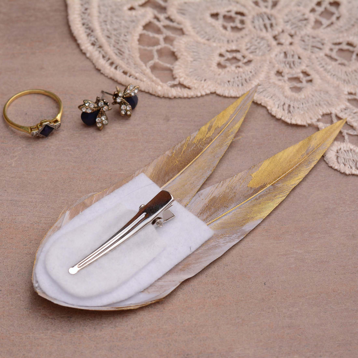A hair clip made from gold feathers on a whit felt backing with a silver toned alligator style hair clip. Displayed on a textured beige surface with a lace doily, a gold ring, and a pair of gold and black stud earrings nearby.