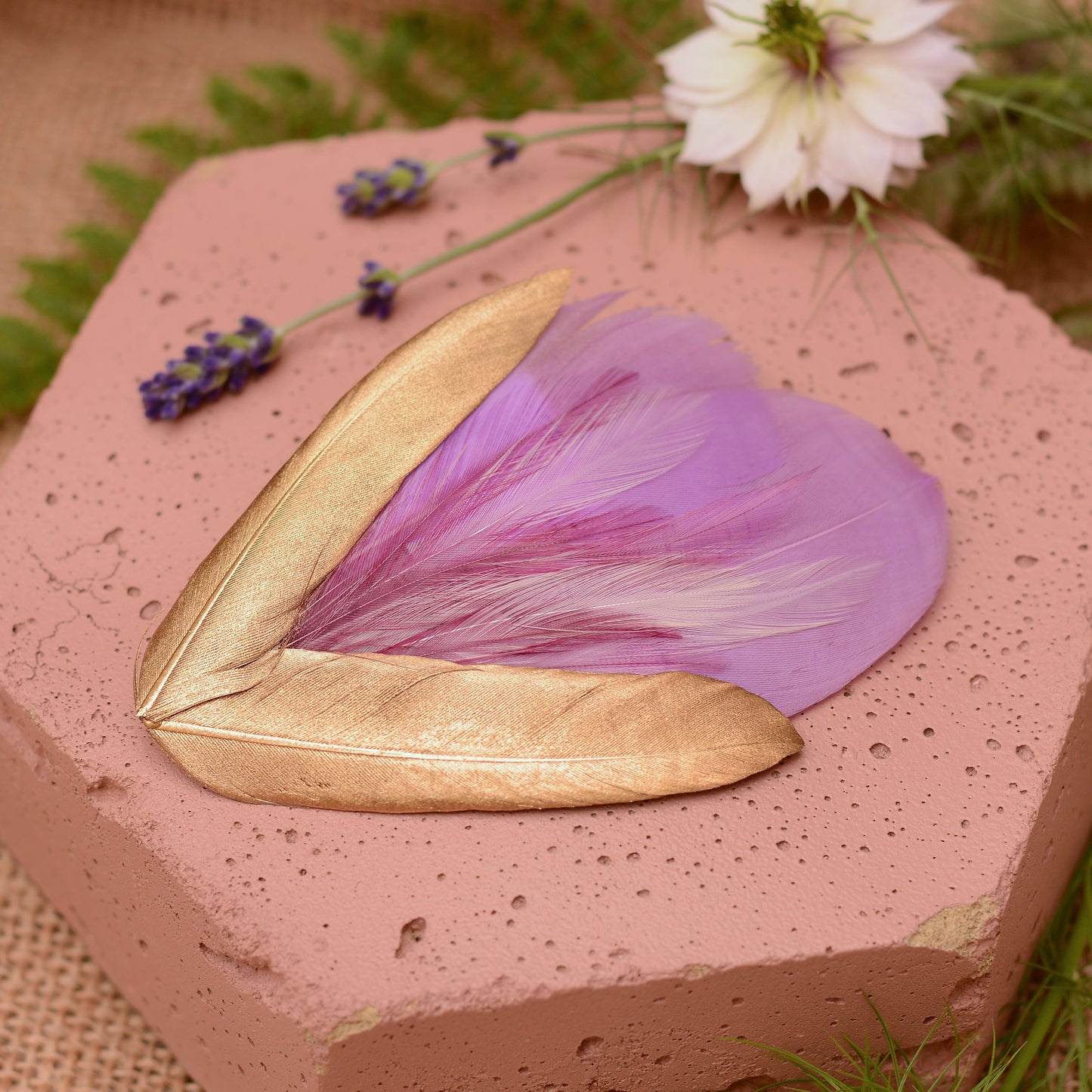 lilac purple and gold decorative feather hair clip fascinator for weddings and special occasions laid on a pink stone surrounded by flowers