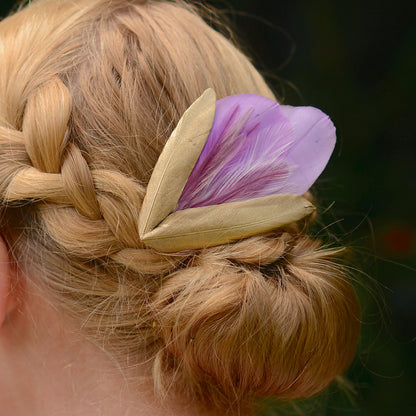 close up side view of lilac purple and gold decorative feather hair clip fascinator for weddings and special occasions laid on a a blonde model with plaited low bun up-do