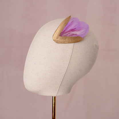 lilac purple and gold decorative feather hair clip fascinator for weddings and special occasions laid the side of an canvas covered mannequin head