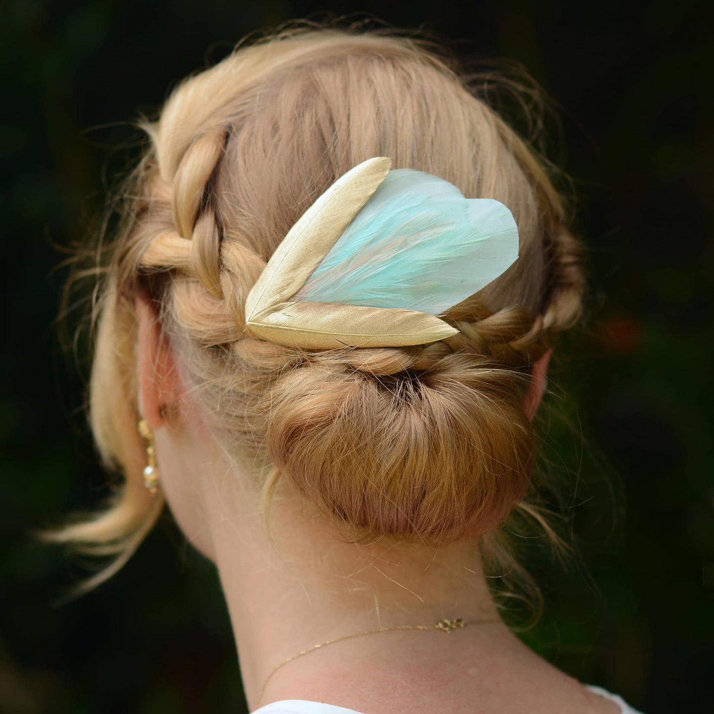 Person with braided hair  and a decorative mint green and gold feather hairpiece against a dark background