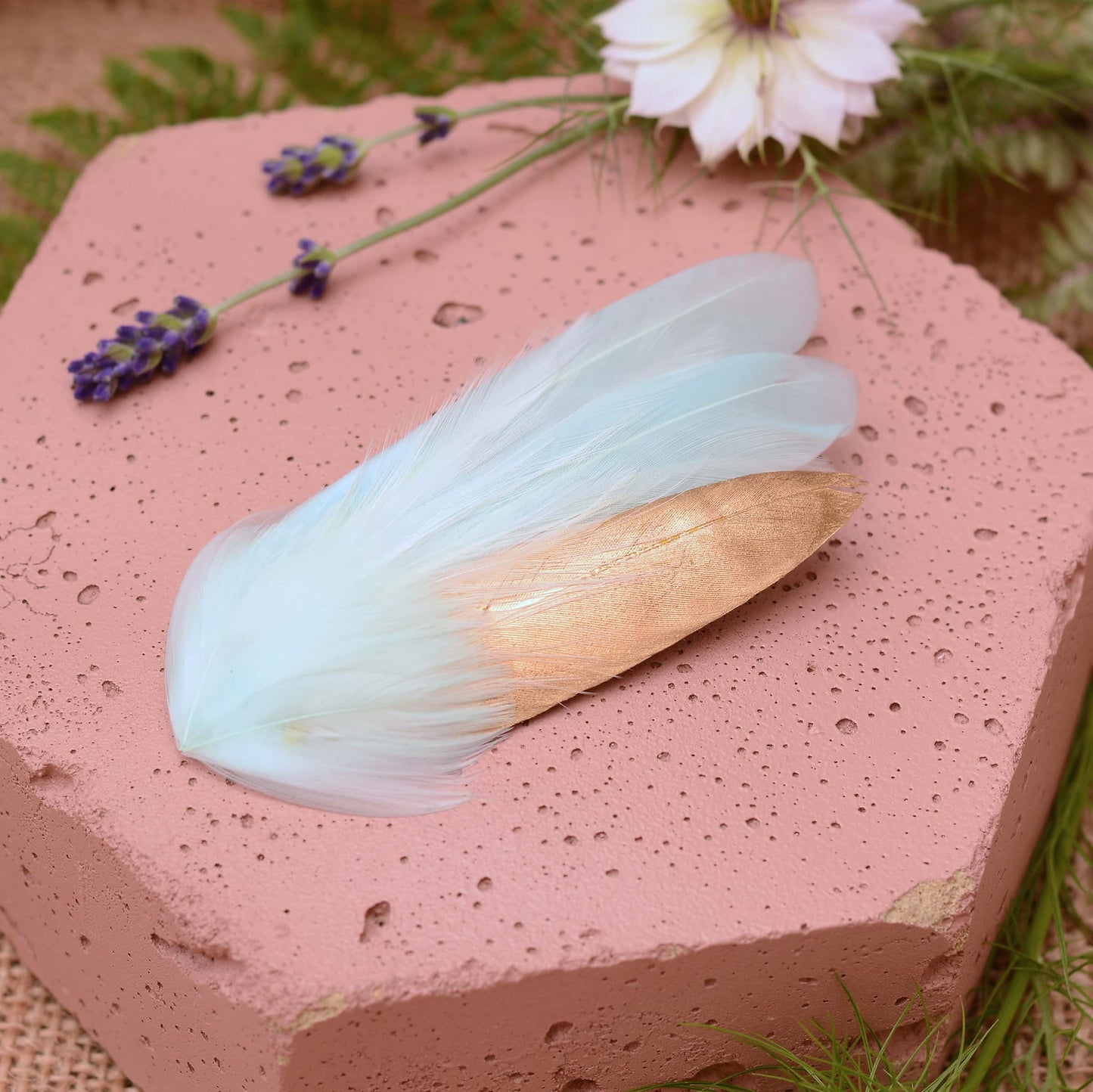 Small mint green and gold feather hair clip on a pink textured stone surrounded with burlap and flowers