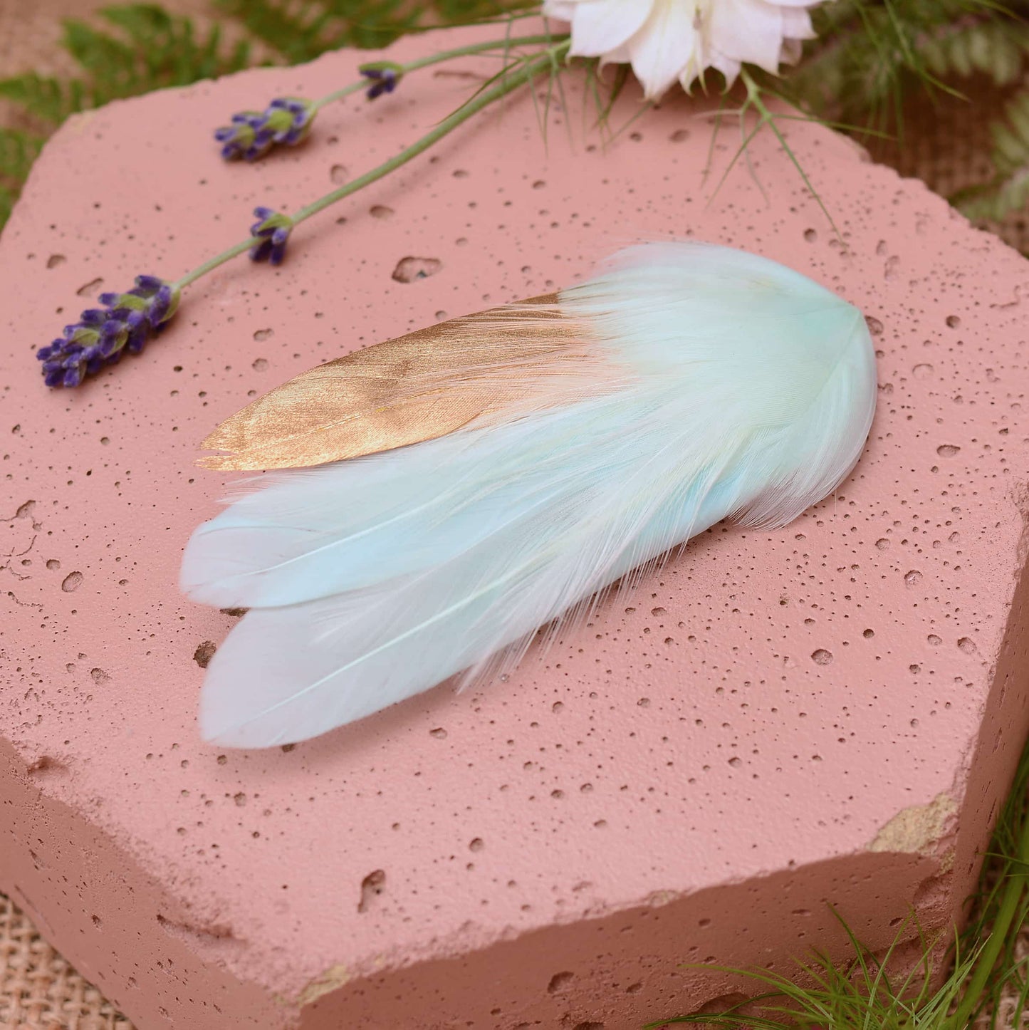 Small mint green and gold feather hair clip on a pink textured stone surrounded with burlap and flowers
