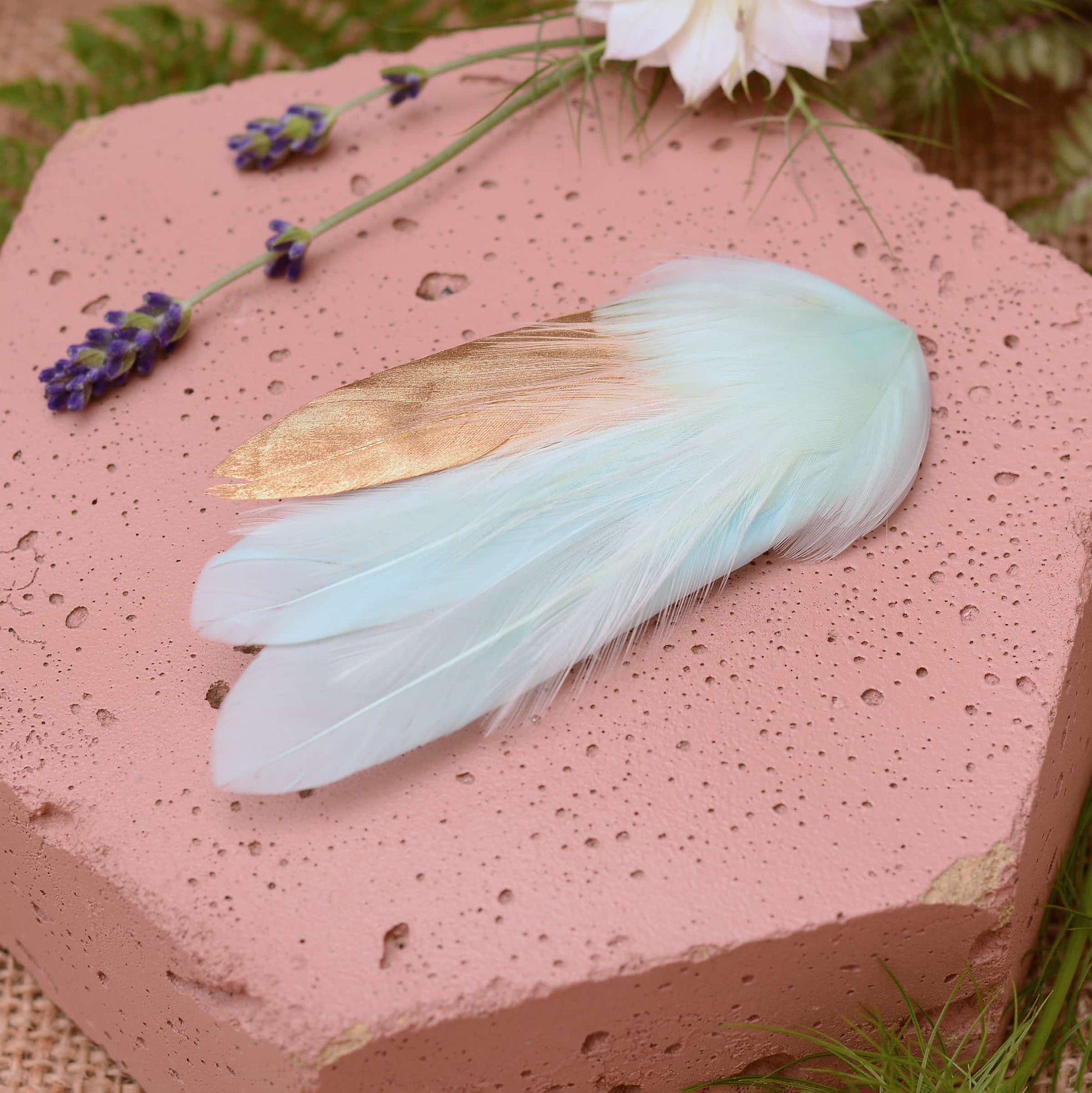 Small mint green and gold feather hair clip on a pink textured stone surrounded with burlap and flowers