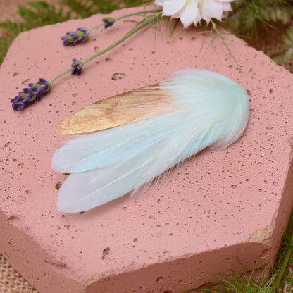 Small mint green and gold feather hair clip on a pink textured stone surrounded with burlap and flowers