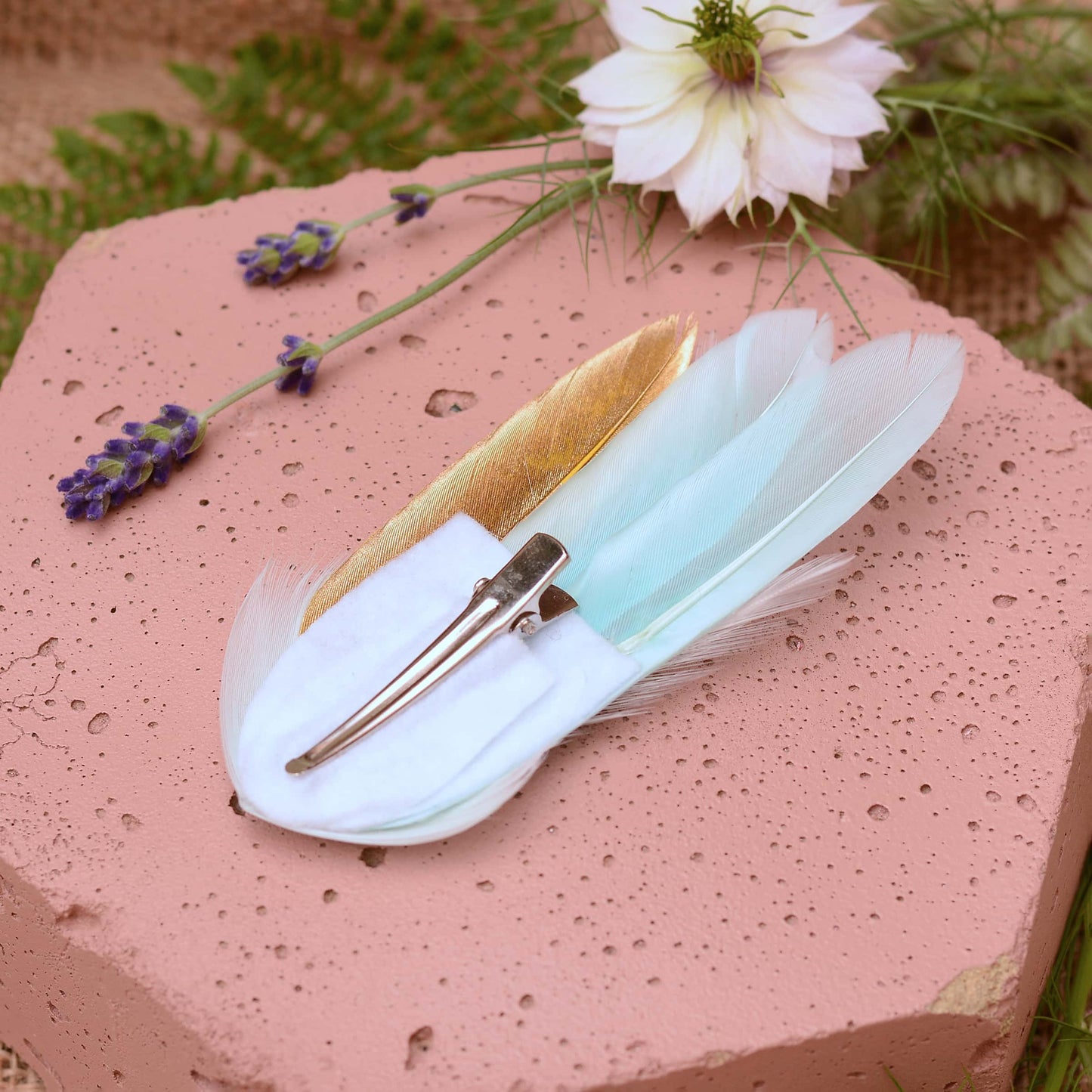 Small mint green and gold feather hair clip on a pink textured stone surrounded with burlap and flowers