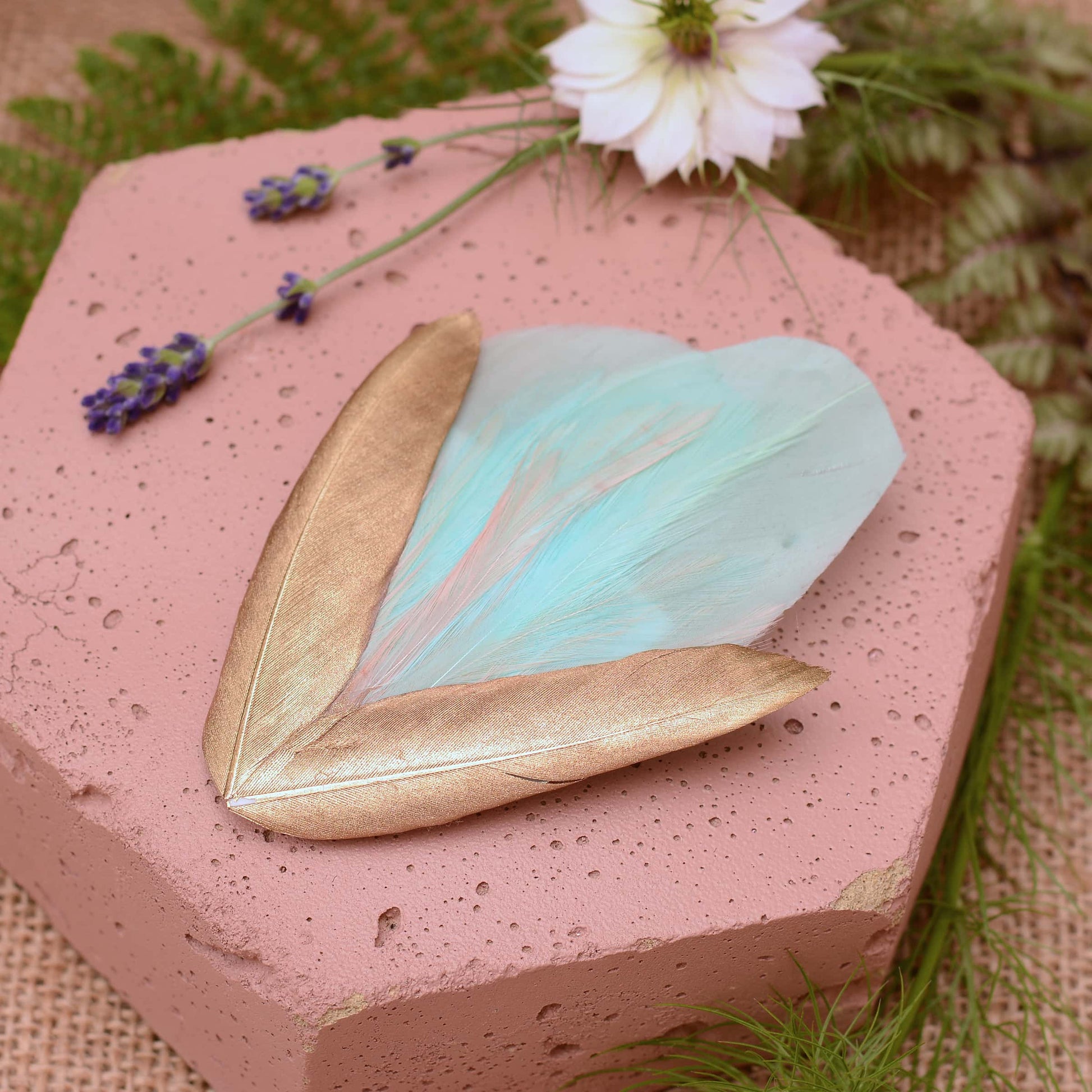 Large mint green and gold feather hair clip on a pink textured stone surrounded with burlap and flowers