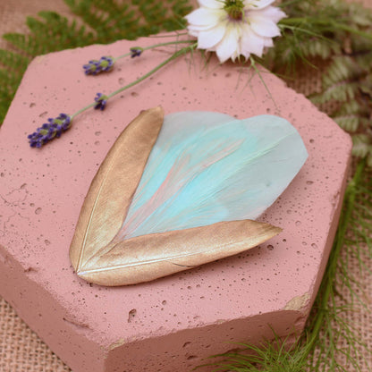 Large mint green and gold feather hair clip on a pink textured stone surrounded with burlap and flowers