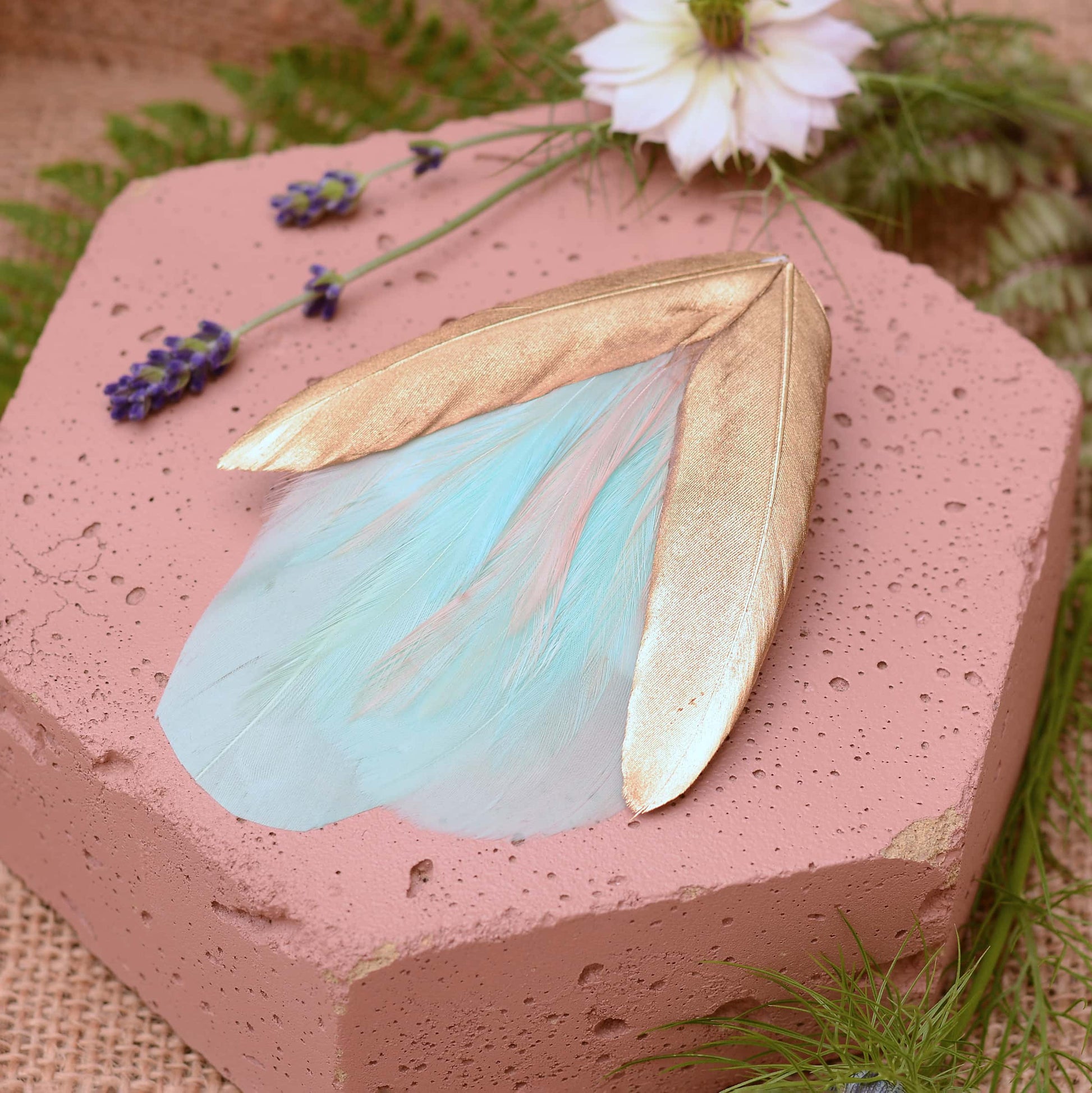 Large mint green and gold feather hair clip on a pink textured stone surrounded with burlap and flowers