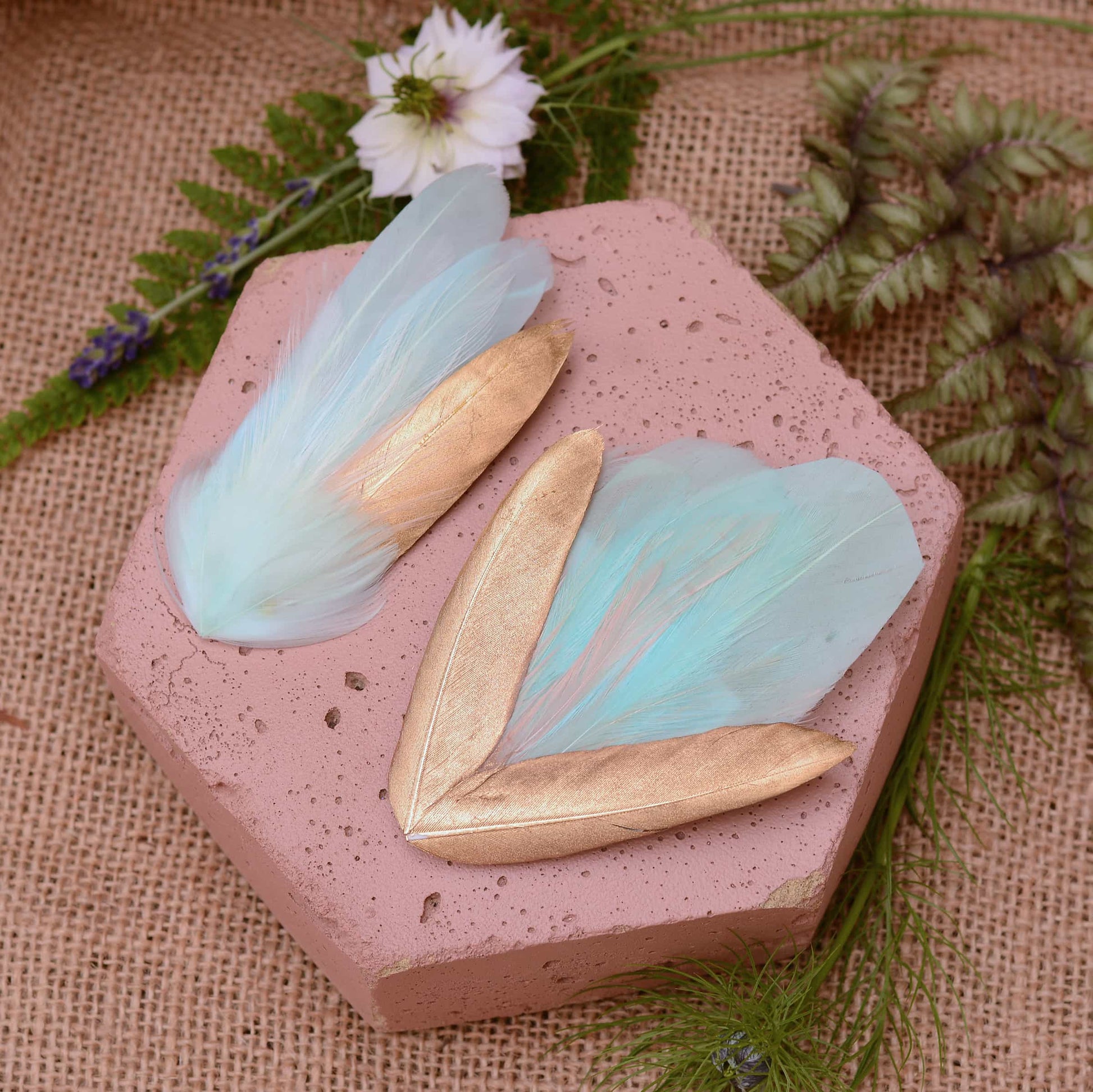 mint green and gold feather hair clips on a pink textured stone surrounded with burlap and flowers