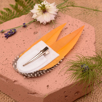 Feathered hair clip with mustard yellow and polka dot  feathers on a pink textured surface with greenery.