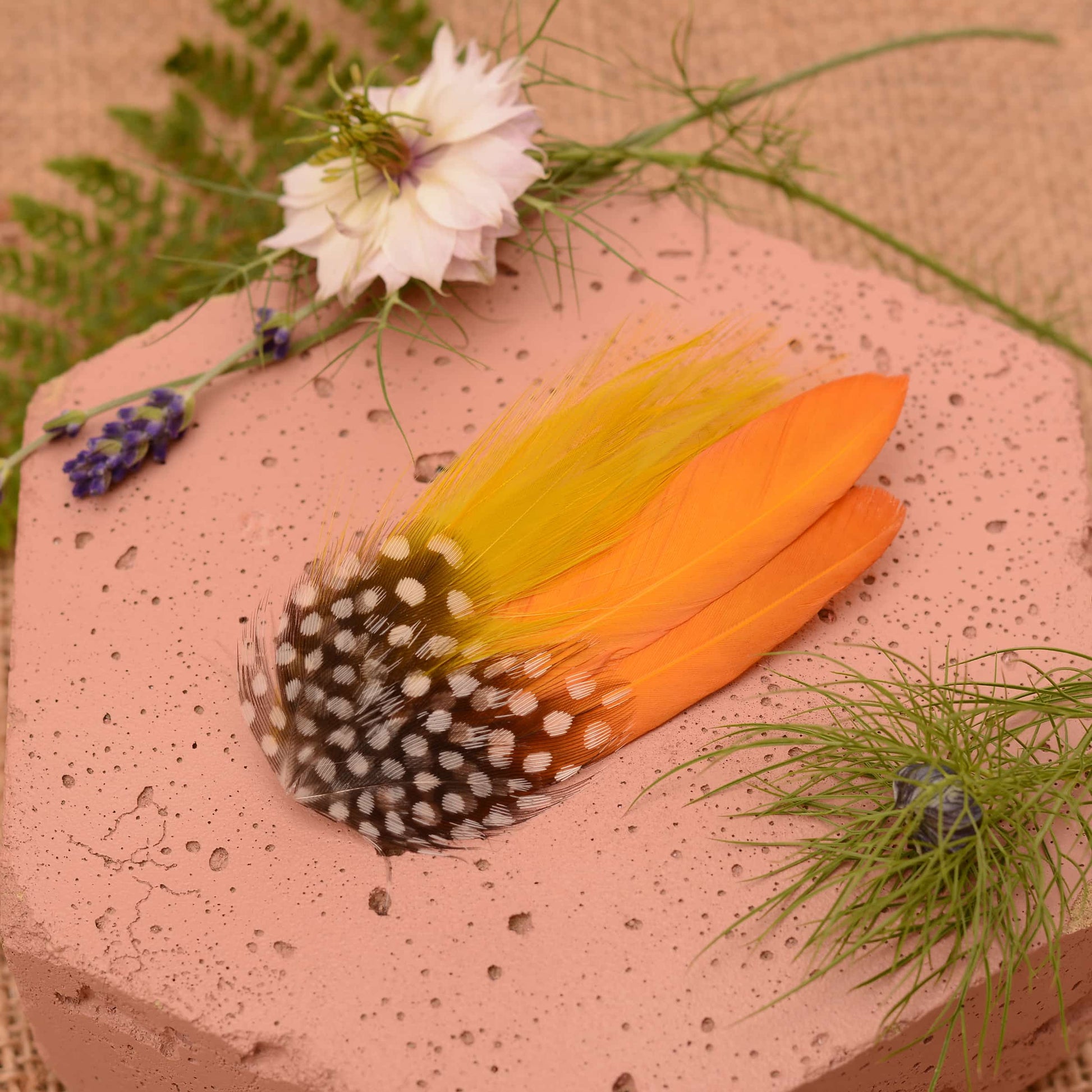 Feathered hair clip with mustard yellow and polka dot  feathers on a pink textured surface with greenery.