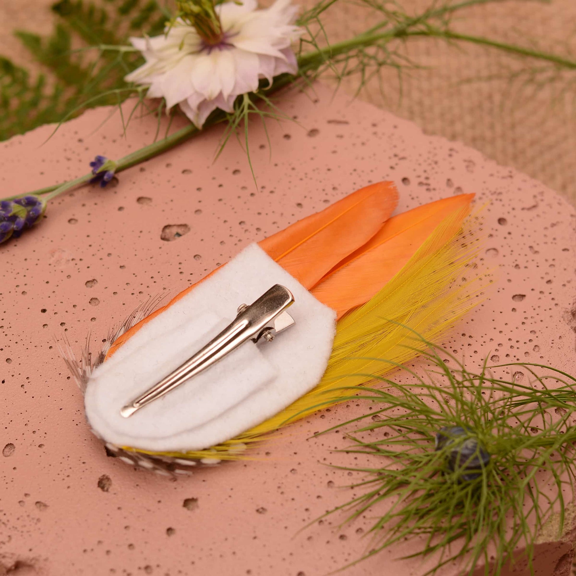 Feathered hair clip with mustard yellow feathers on a pink textured surface with greenery.