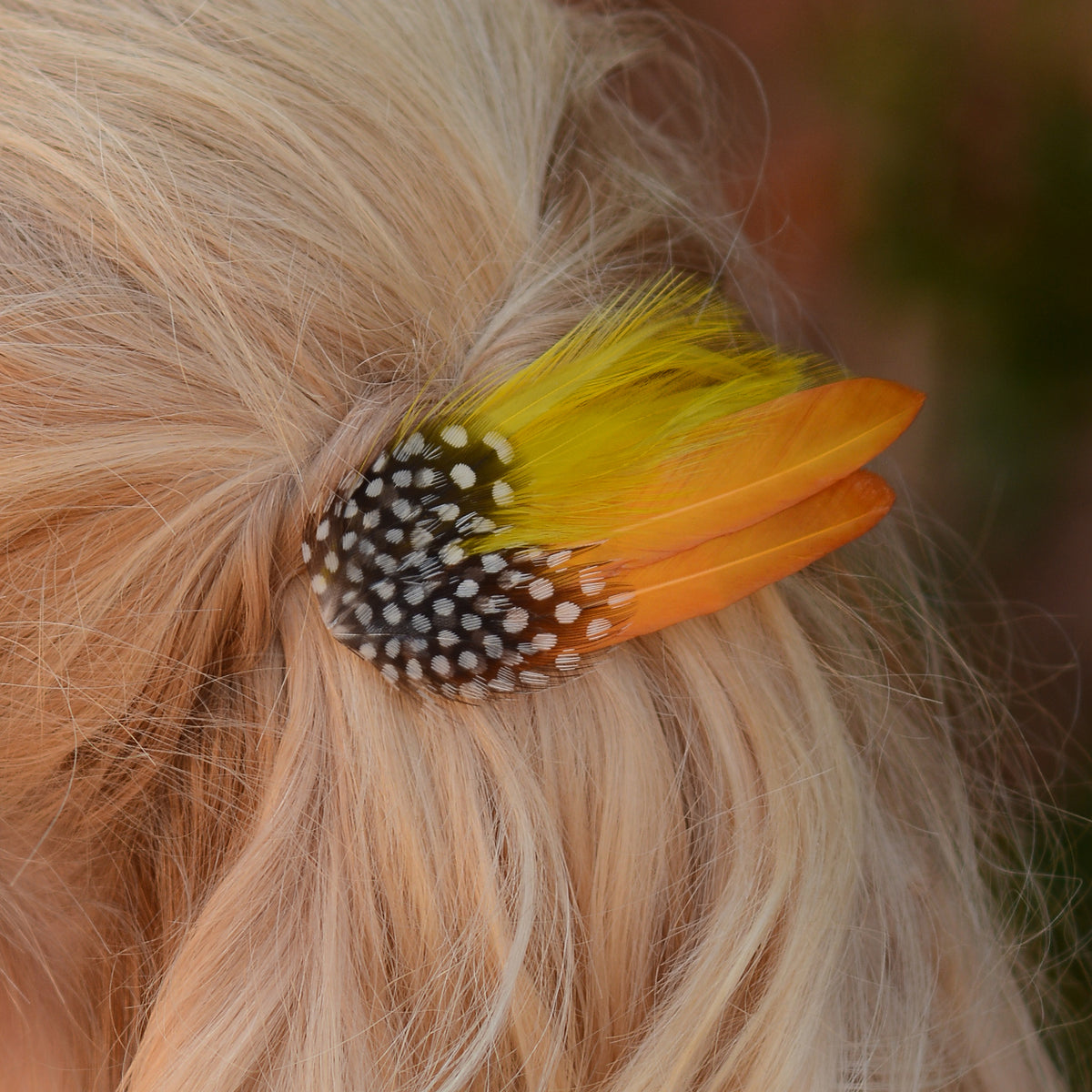 A close-up of blonde hair secured with a decorative feather hair clip featuring golden mustard yellow feathers and black and white polka dot feathers.