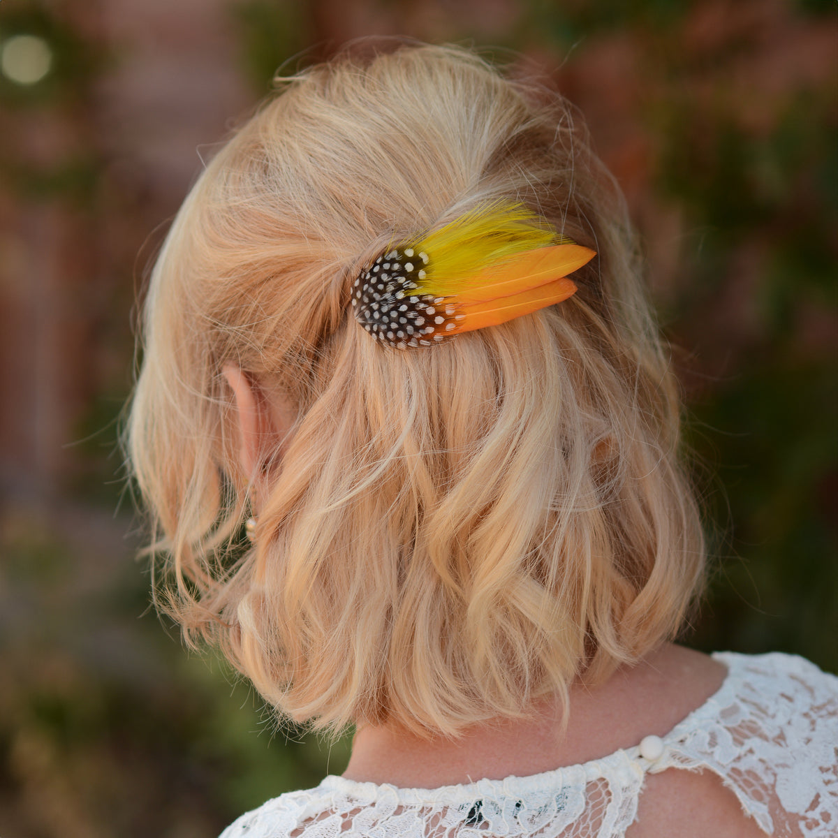 A blonde woman shown from the back wearing a decorative hair claw clip with golden mustard yellow feathers and a black-and-white spotted feathers, set against a blurred outdoor background.
