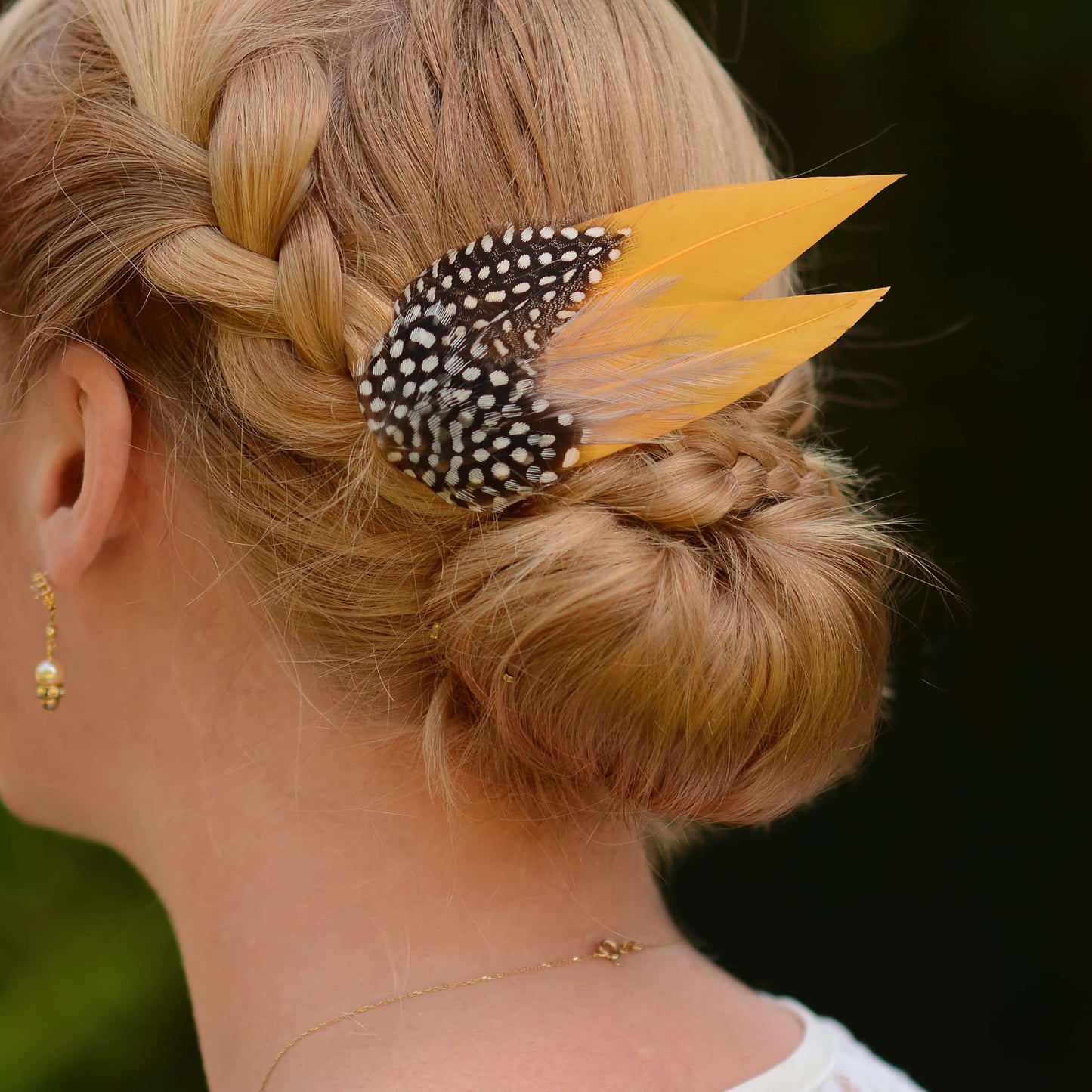 Woman with braided hair wearing a decorative feather hair clip ain gold yellow and polka dot feathers against a dark background