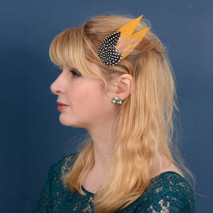 Woman with a feathered hair clip in mustard yellow and polka dot  feathers against a blue background