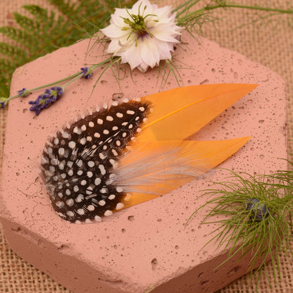 Feathered hair clip with mustard yellow and polka dot  feathers on a pink textured surface with greenery.