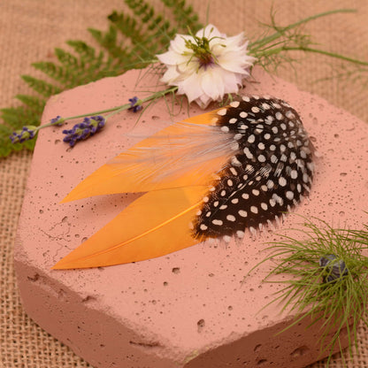 Feathered hair clip with mustard yellow and polka dot  feathers on a pink textured surface with greenery.
