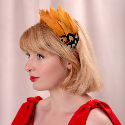 A woman in profile wearing a bright gold yellow feather headband with a brown, black and white patterned feathers, paired with a red dress against a soft beige curtain background.