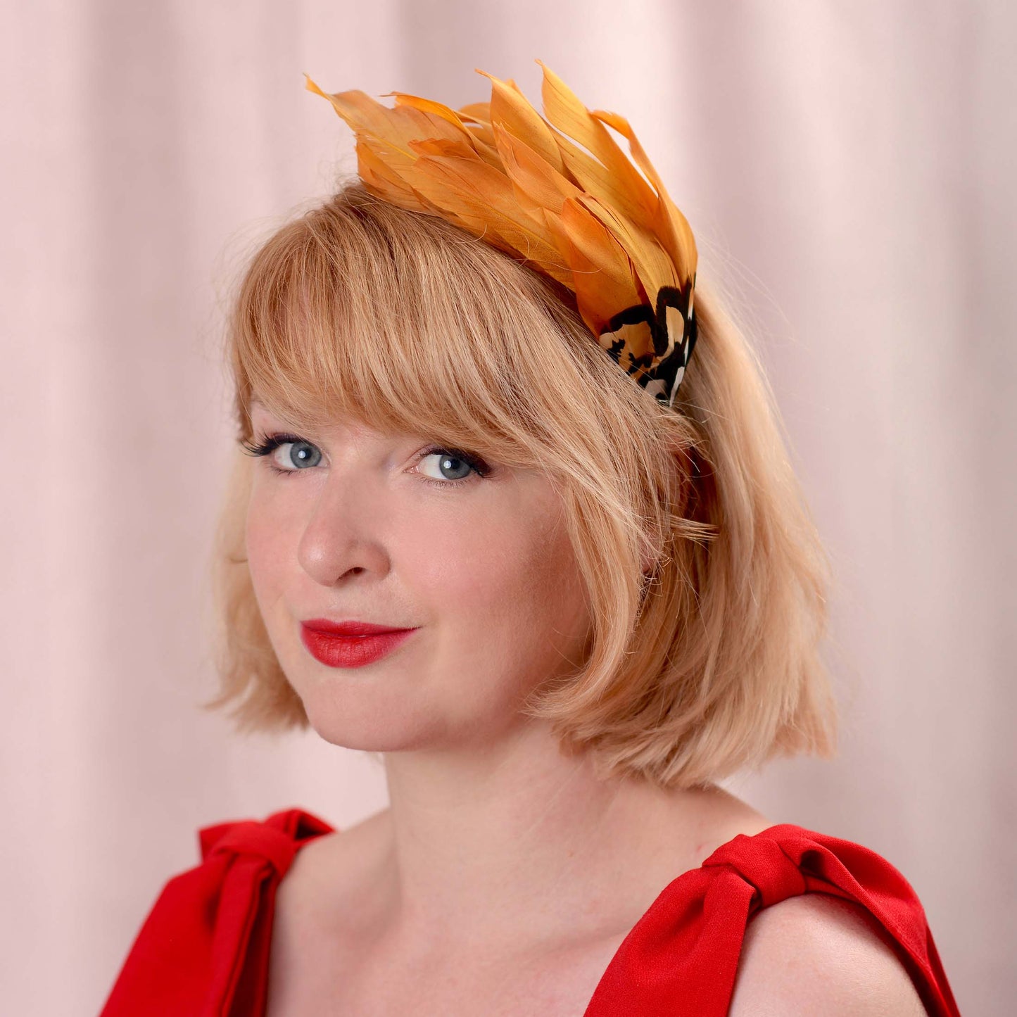 A woman in three quarter view wearing a bright gold yellow feather headband with a brown, black and white patterned feathers, paired with a red dress against a soft beige curtain background.