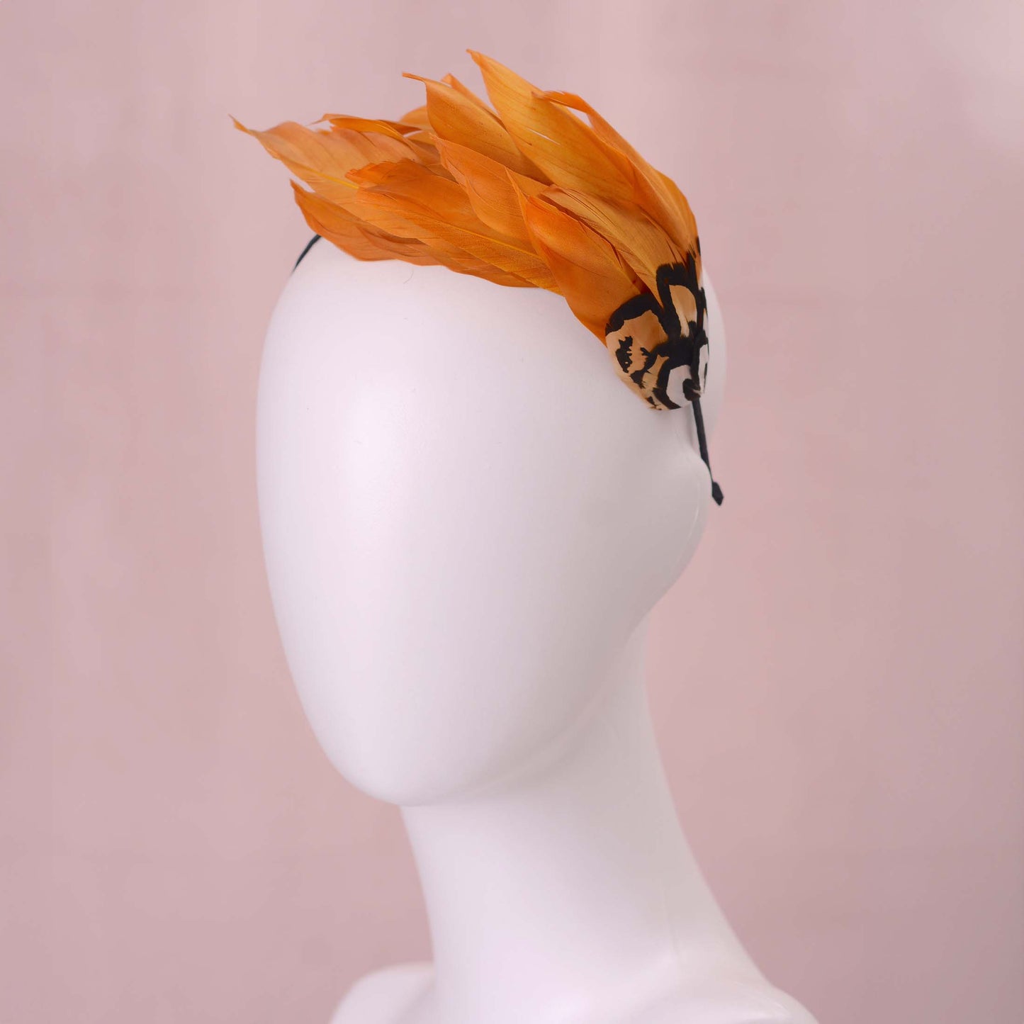 A gold yellow feather fascinator headpiece with brown, black and gold patterned feathers displayed on a white mannequin head against a soft pink background.