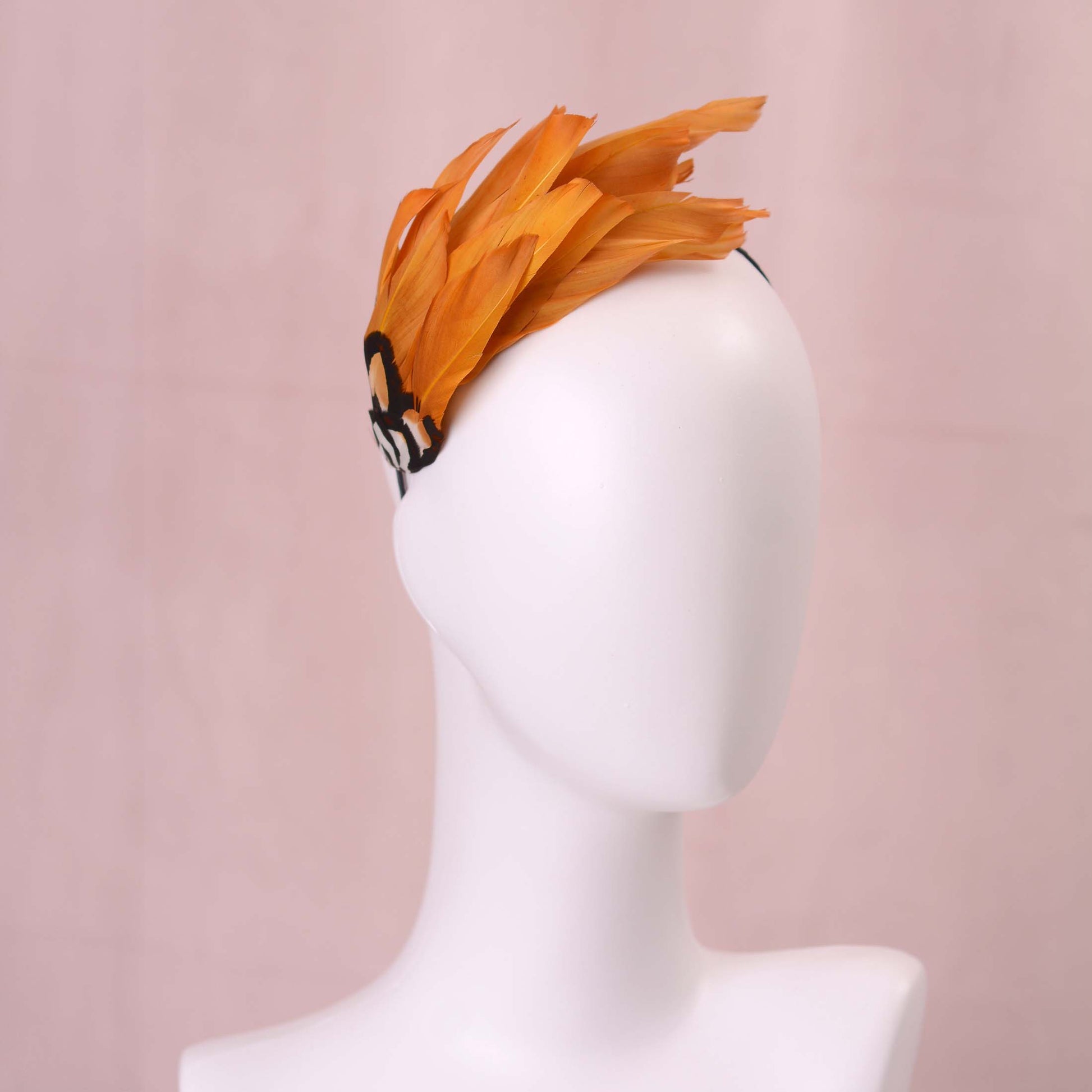 A gold yellow feather fascinator headpiece with brown, black and gold patterned feathers displayed on a white mannequin head against a soft pink background.