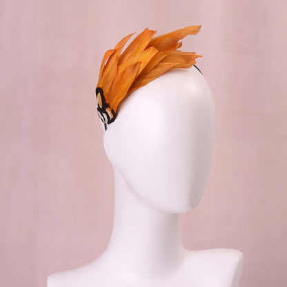 A gold yellow feather fascinator headpiece with brown, black and gold patterned feathers displayed on a white mannequin head against a soft pink background.