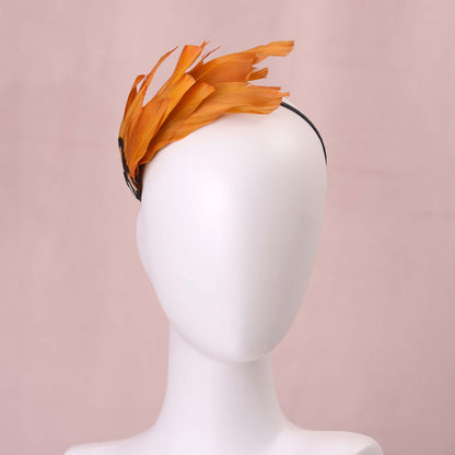 A gold yellow feather fascinator headpiece with brown, black and gold patterned feathers displayed on a white mannequin head against a soft pink background.