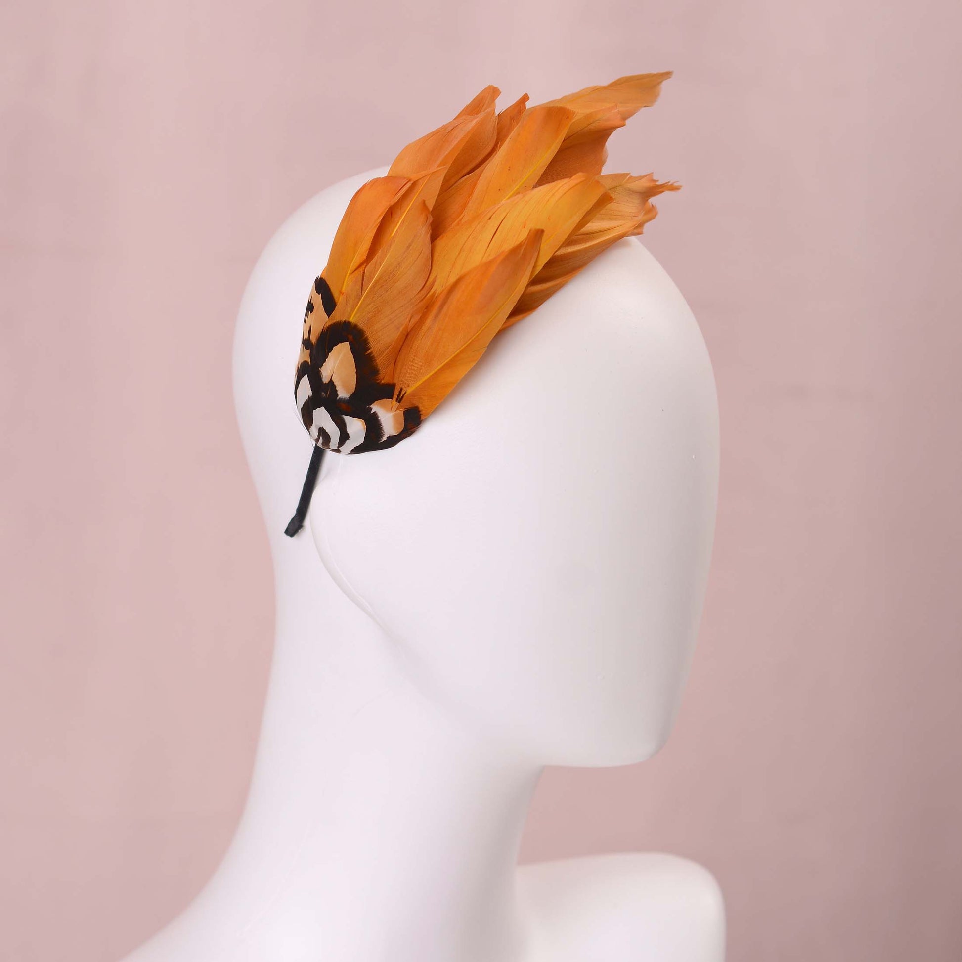 A gold yellow feather fascinator headpiece with brown, black and gold patterned feathers displayed on a white mannequin head against a soft pink background.